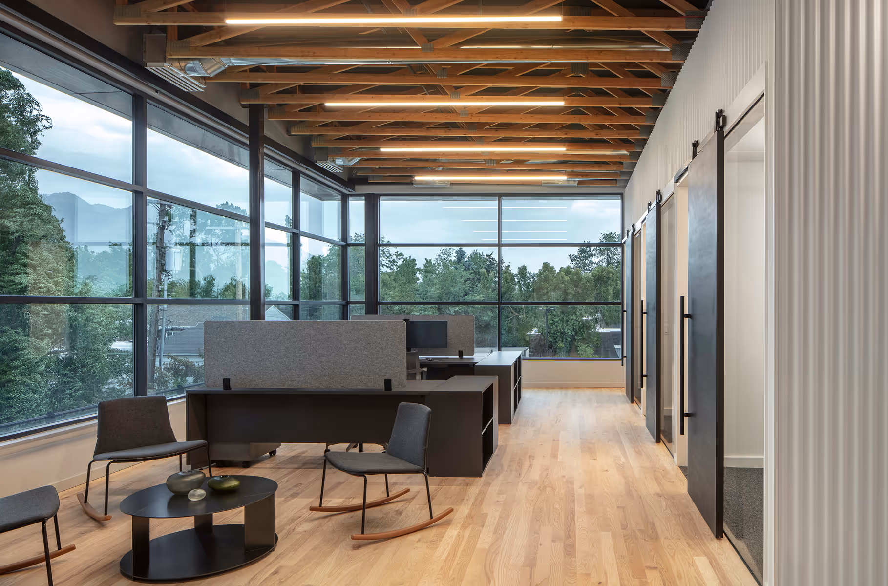 Modern office with wood floor, large windows, desks, chairs, and sliding black doors to rooms.