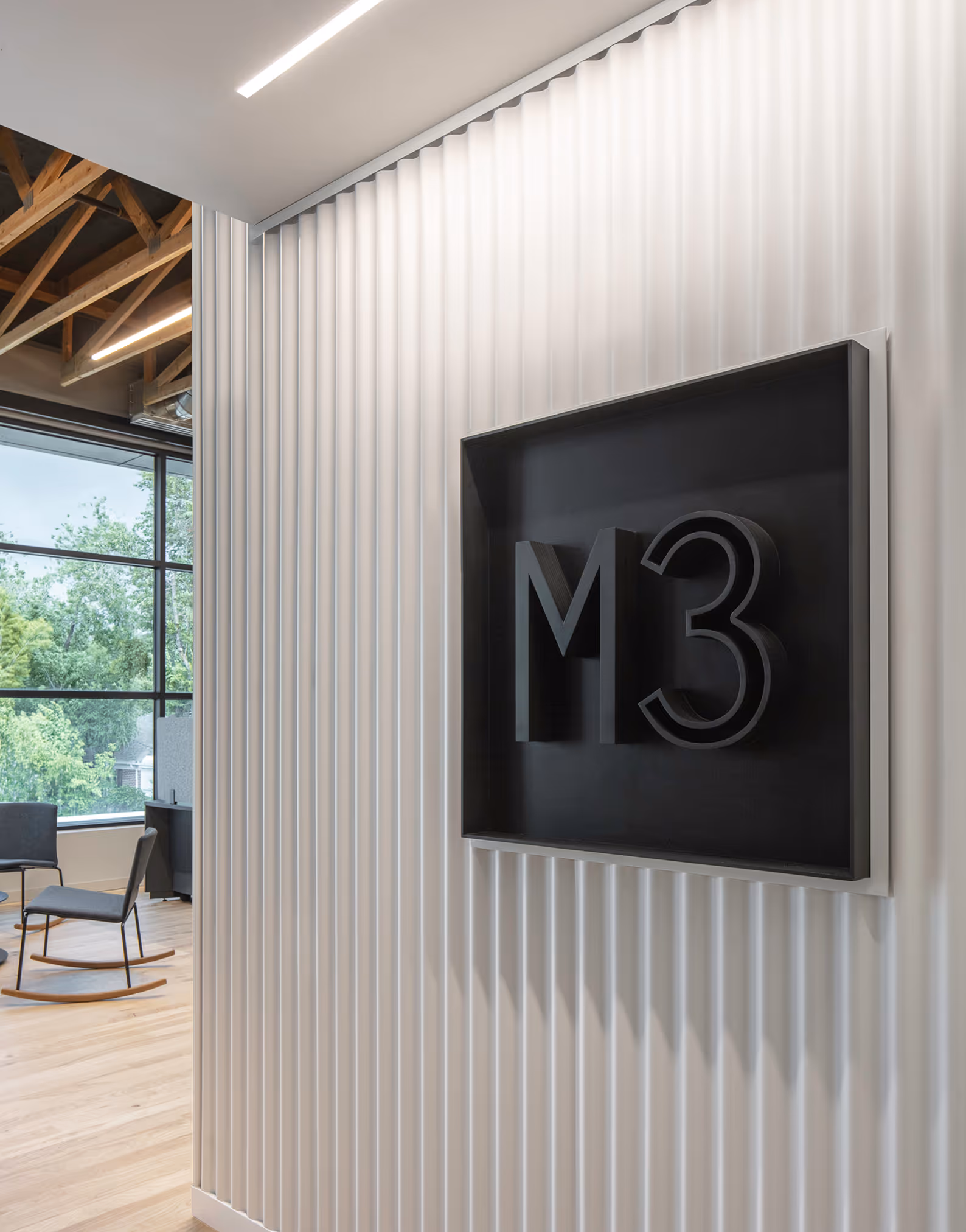 Modern office interior with light wood floor, large window, rocking chairs, and black M3 logo on wall.