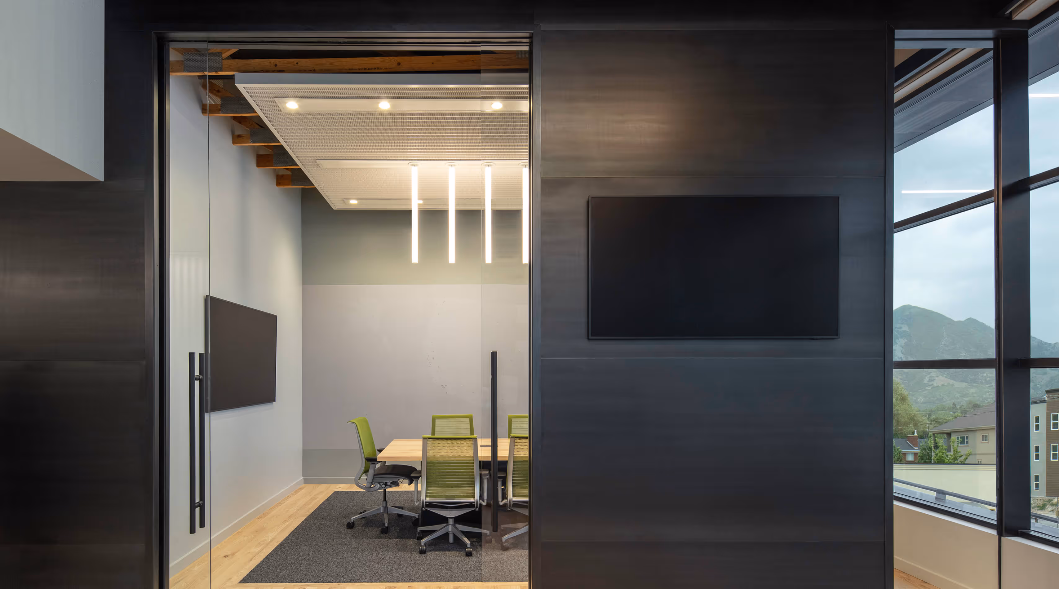 Modern office meeting room with green chairs, wooden table, glass walls, and two wall-mounted TVs.