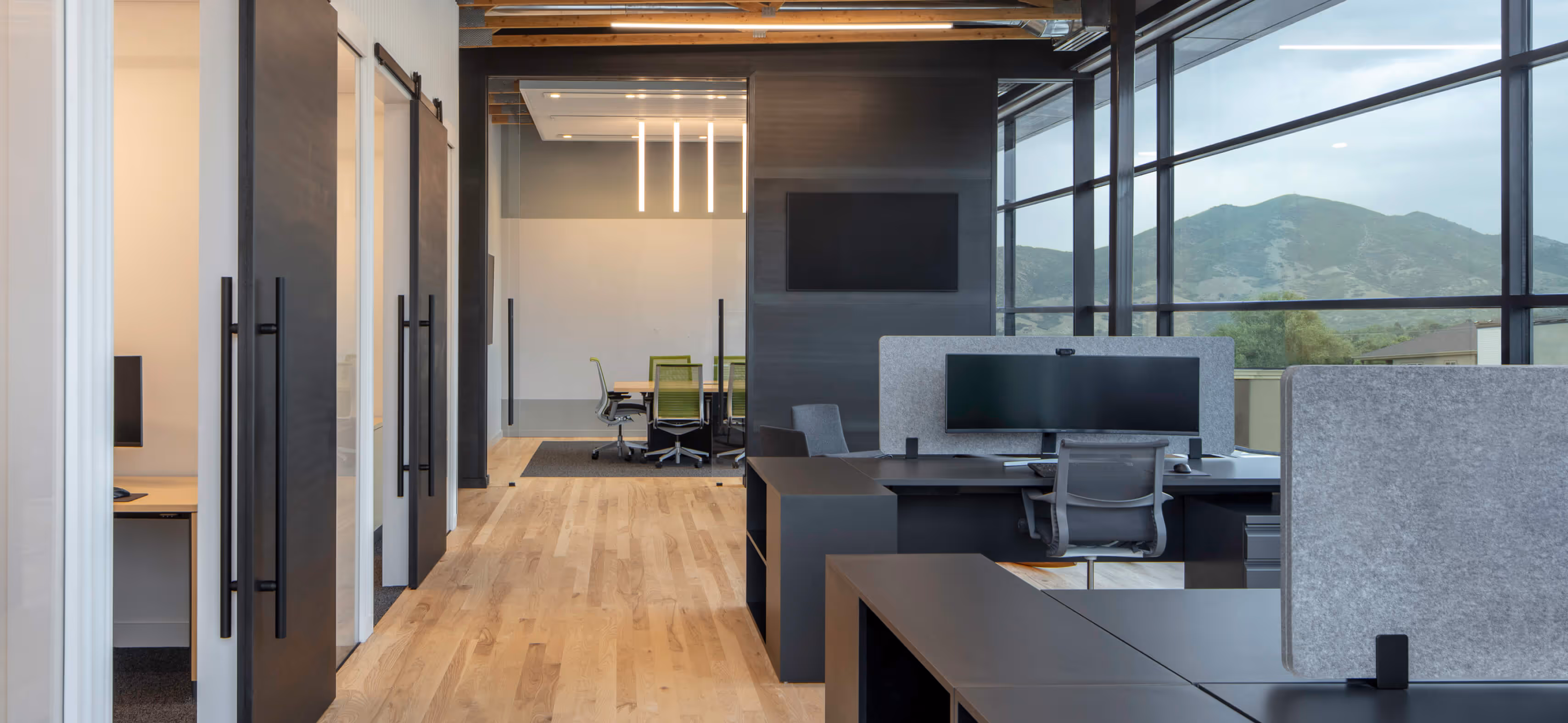 Modern office interior with wooden floors, desks, computers, glass walls, and mountain view outside.
