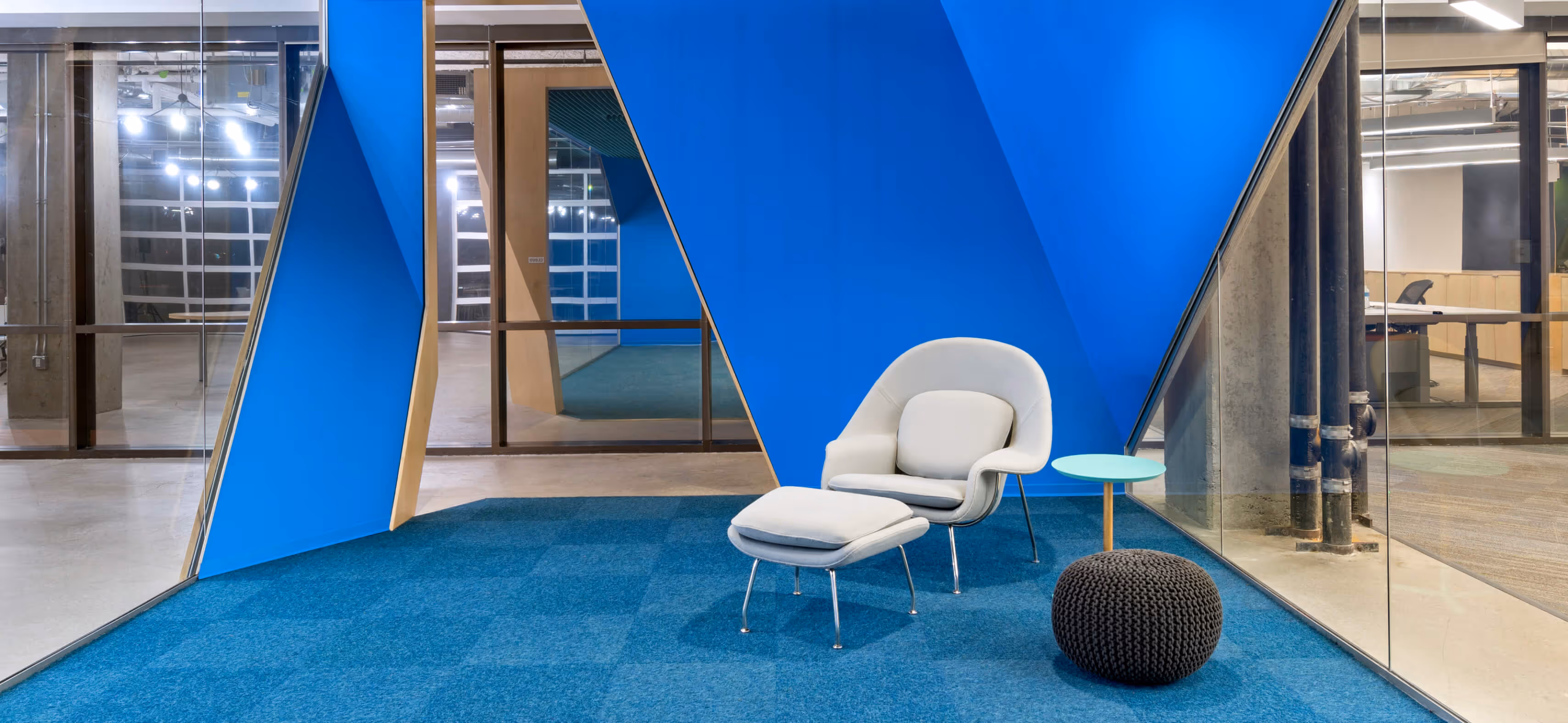 Modern office corner with a light gray lounge chair, matching ottoman, small blue table, and knitted pouf.