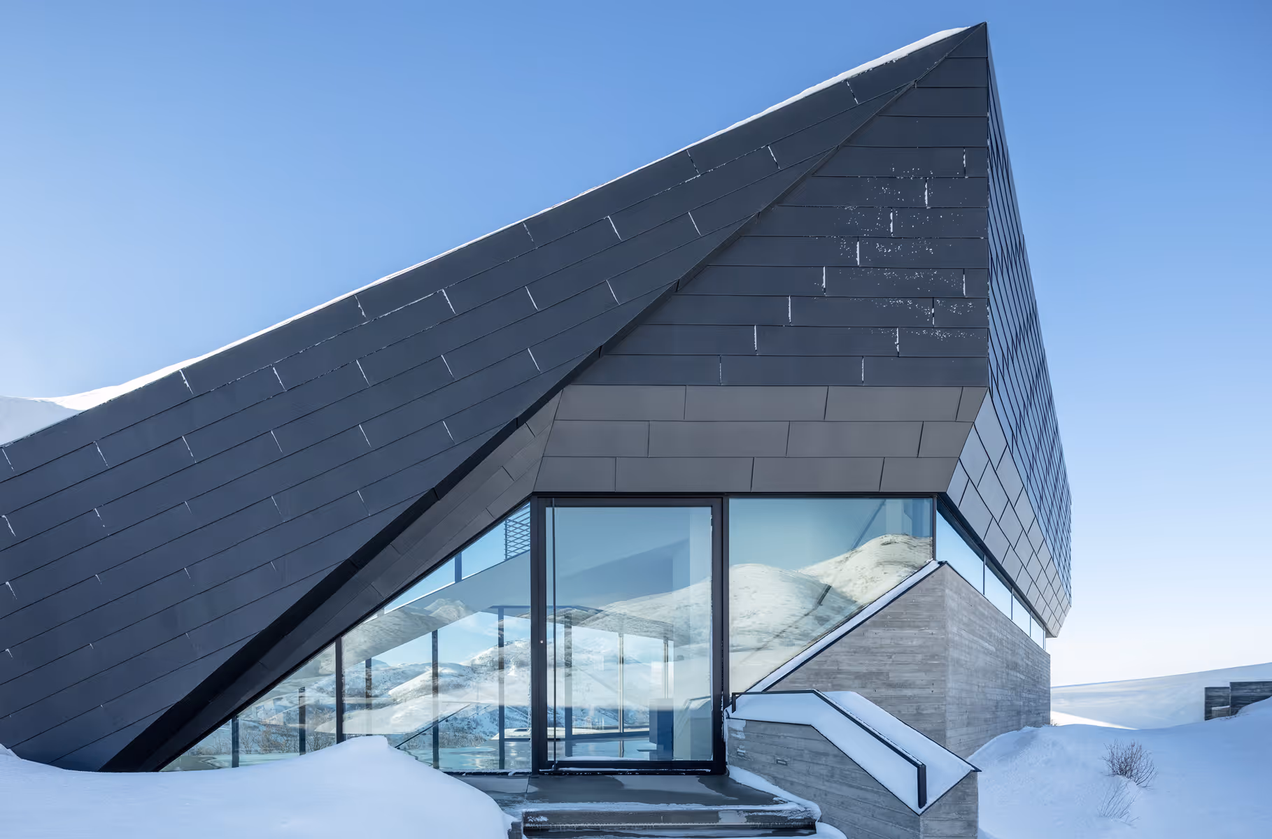 Modern angular house with glass walls and snow-covered roof in a snowy landscape.