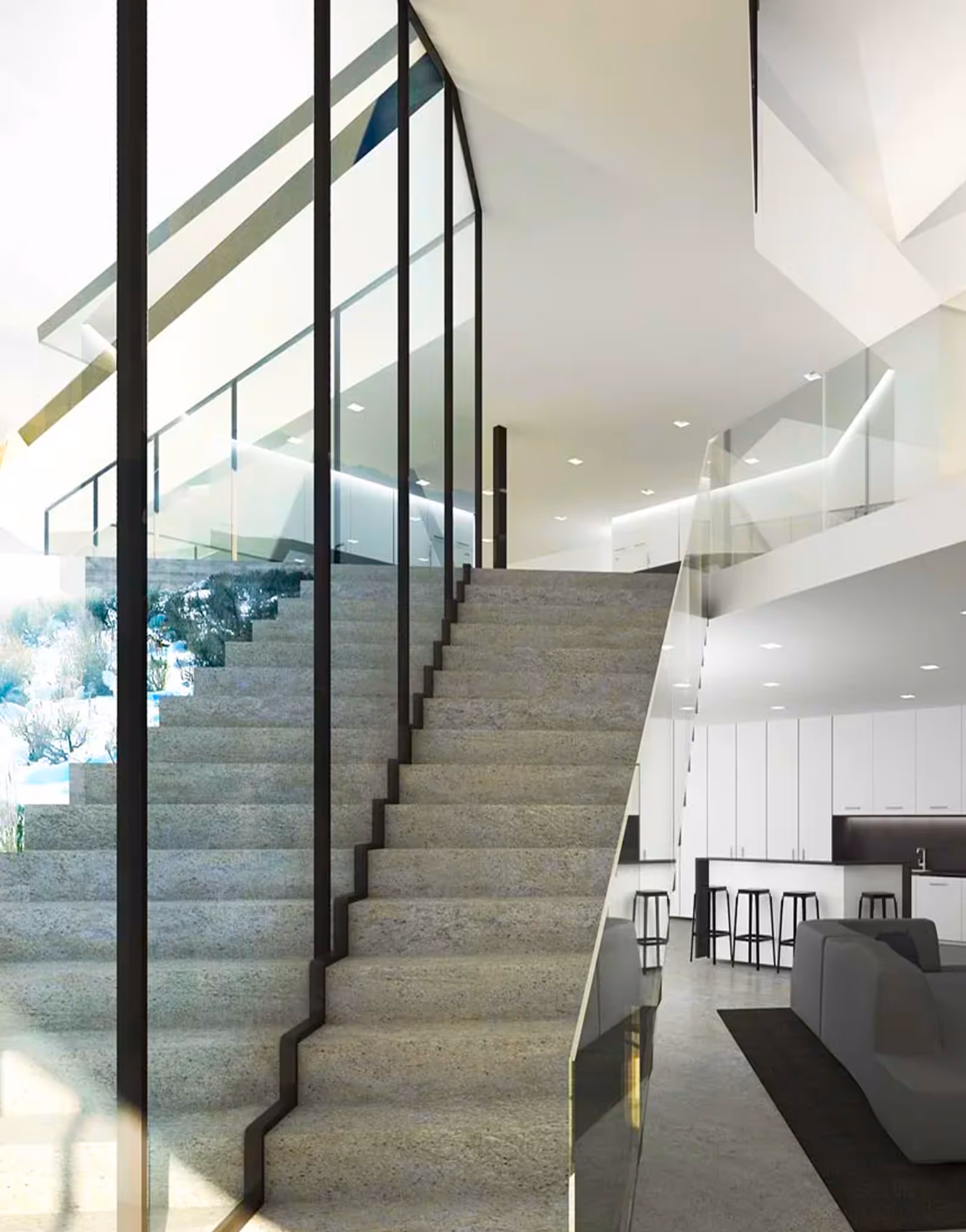 Modern interior with gray stairs, glass railings, white kitchen, black stools, and gray sofas.