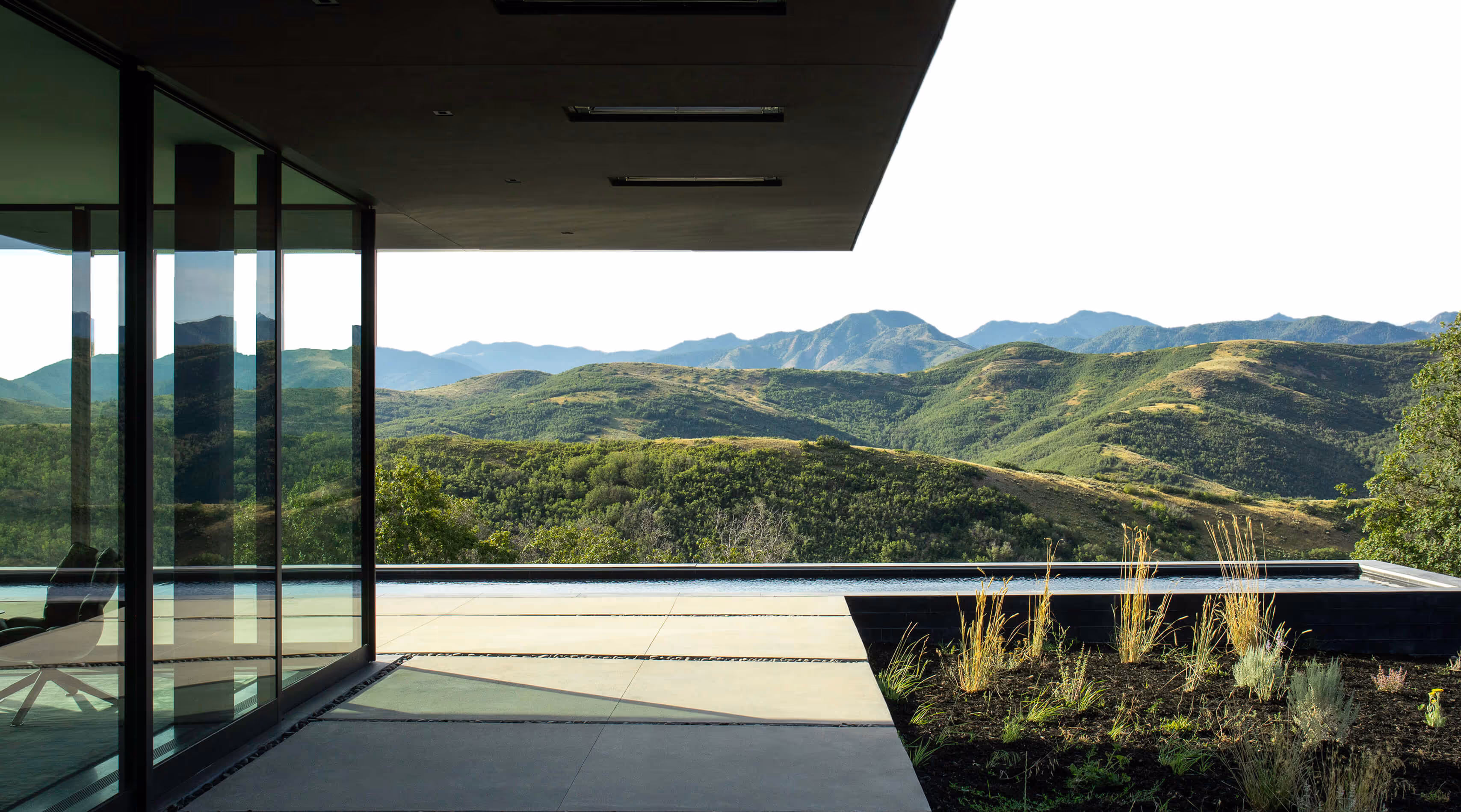 Modern house patio with glass walls, infinity pool, and a scenic view of green rolling hills and mountains.
