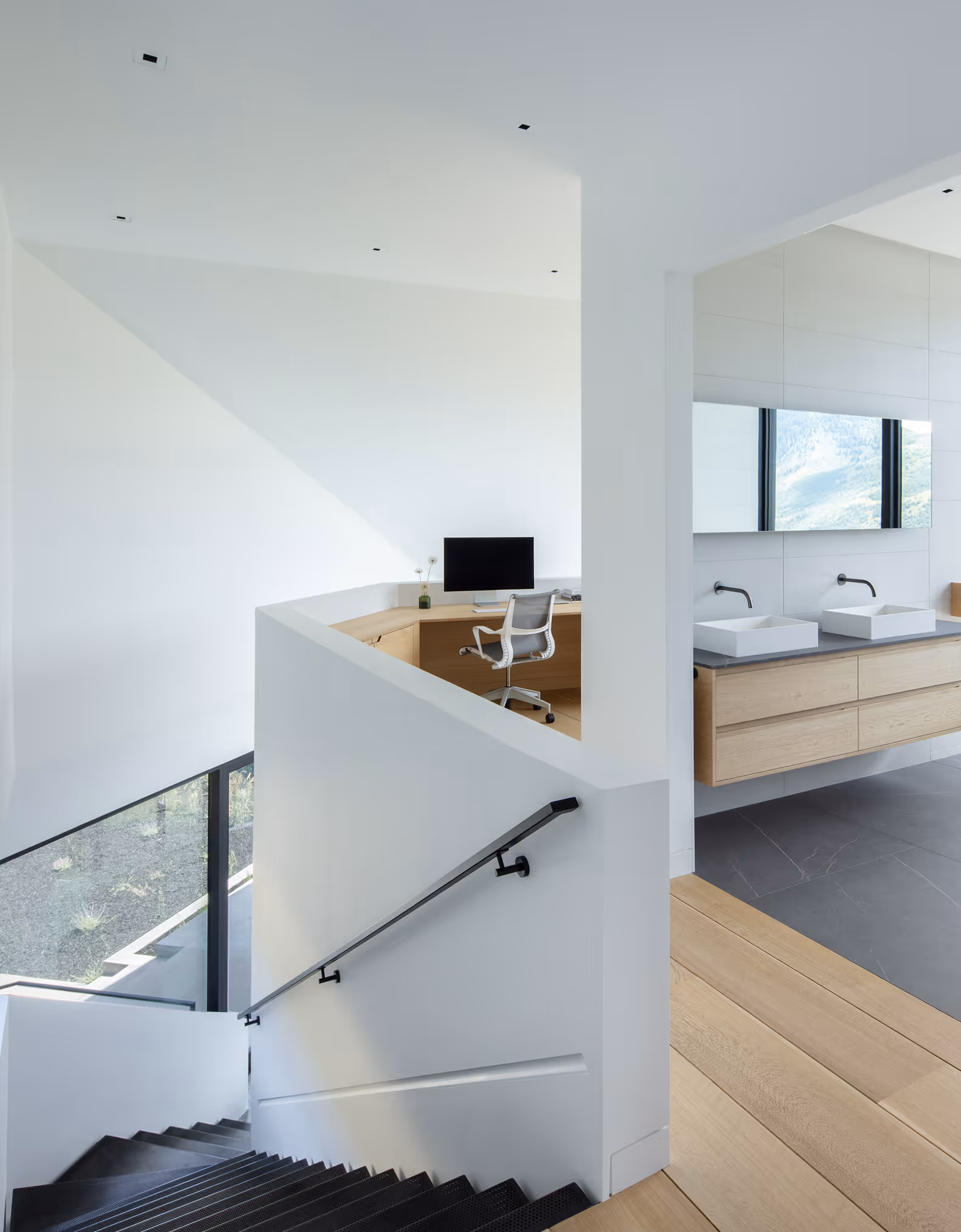 Modern white interior with black stairs, wooden desk with computer, and bathroom with double sinks.