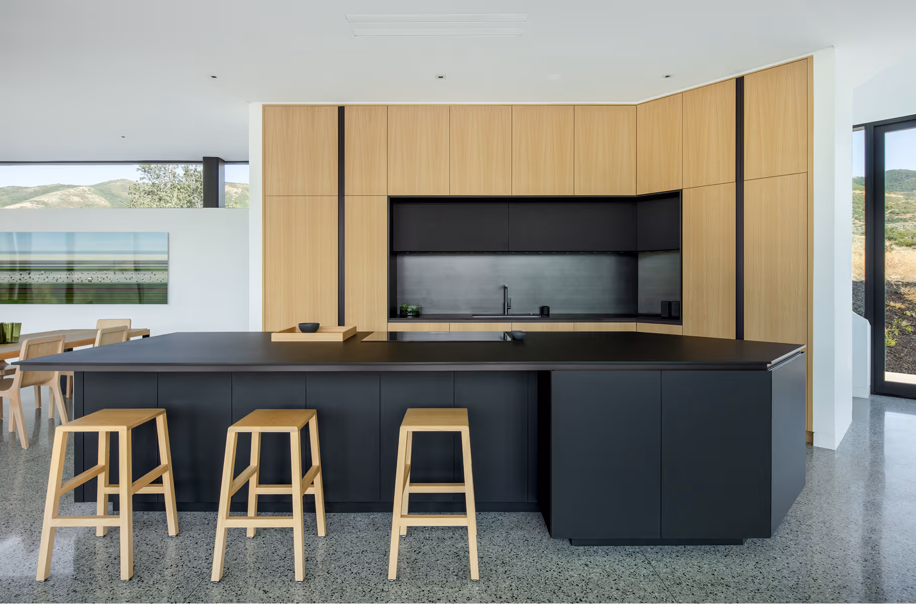 Modern kitchen with black island, light wood cabinets, three wooden stools, and scenic windows.
