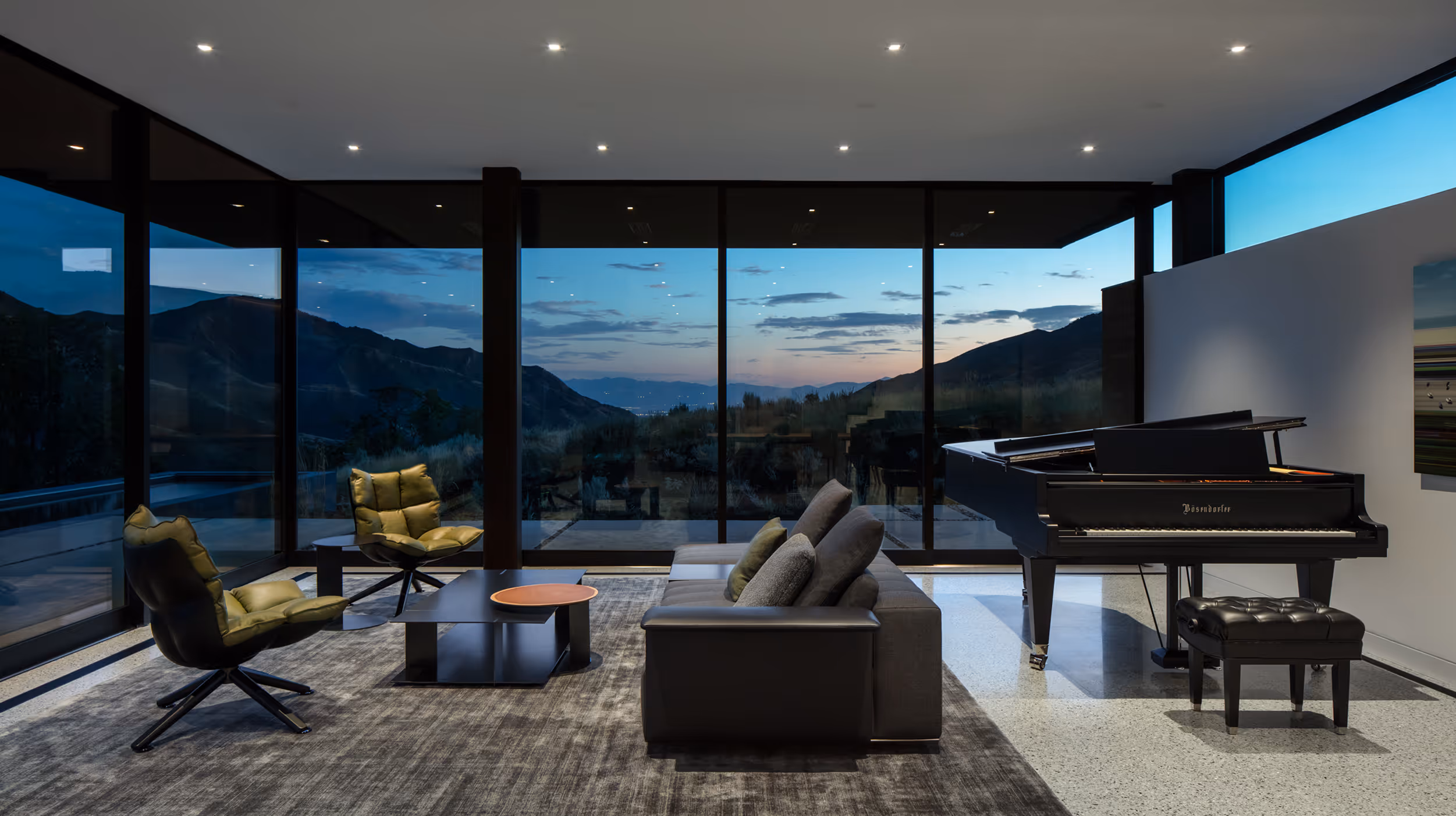 Modern living room with large glass windows, two green chairs, a sofa, coffee table, and grand piano at dusk.