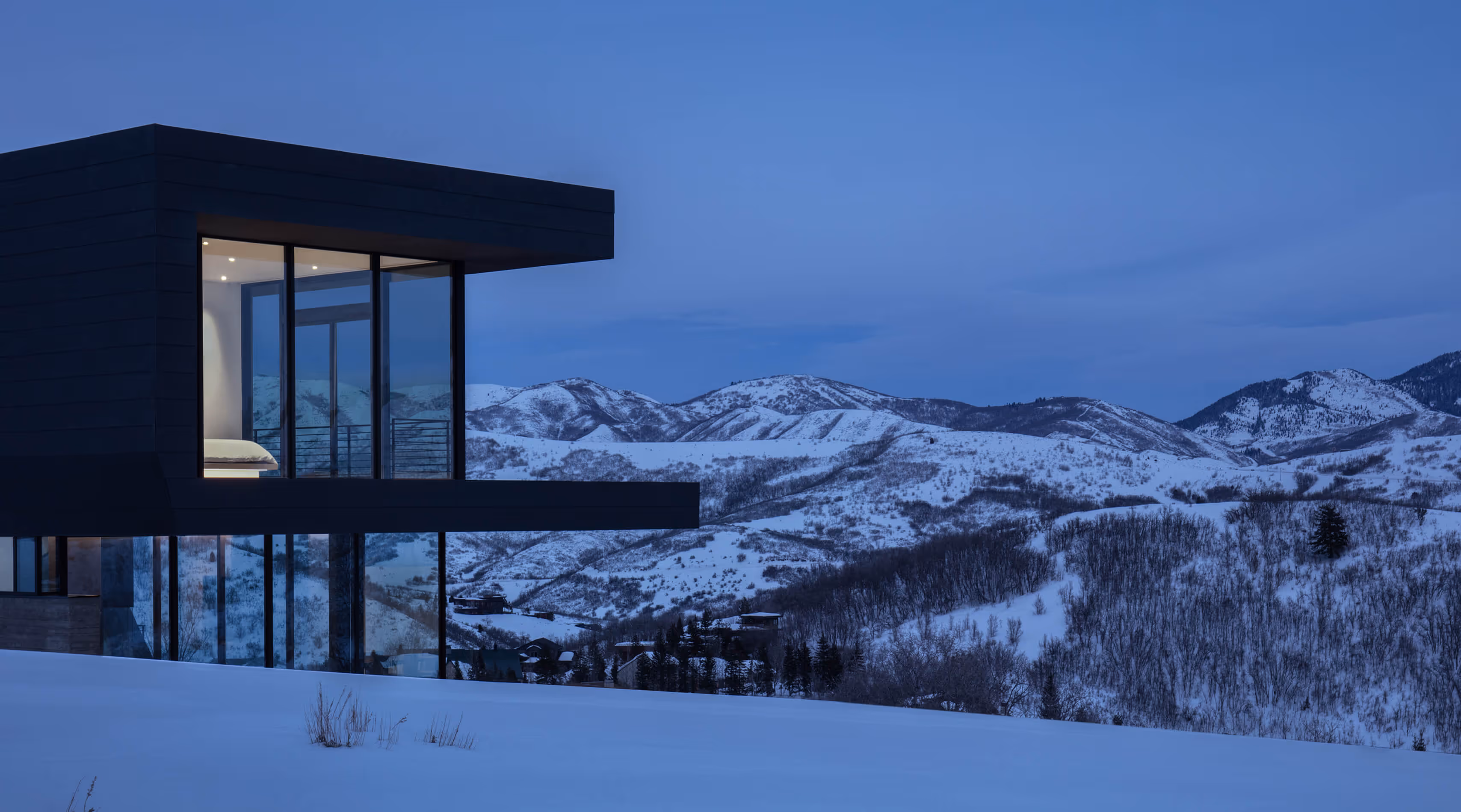 Modern house with large glass windows overlooking snow-covered mountains at dusk.