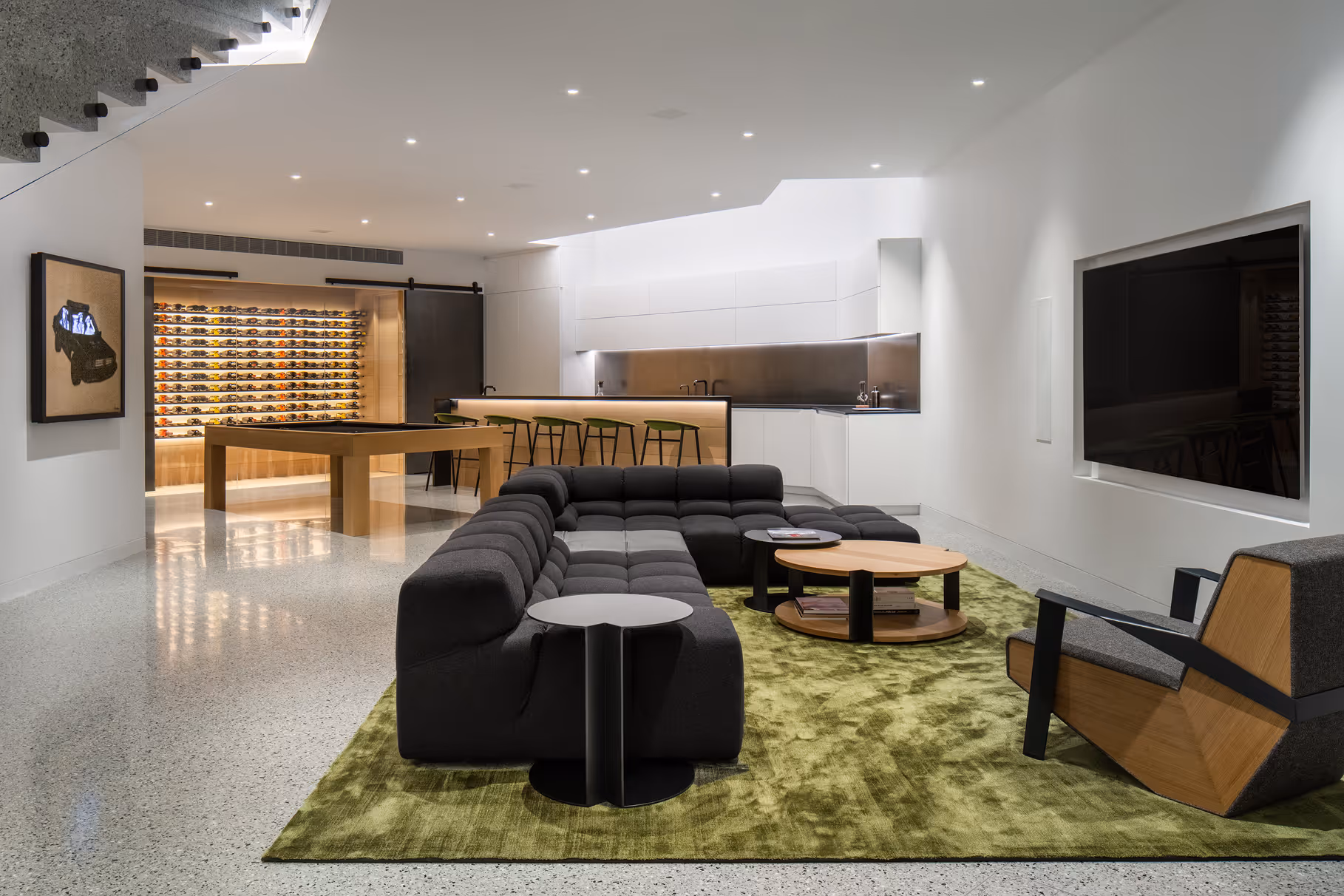 Modern living room with gray sectional sofa on green rug, wood armchair, wall TV, and illuminated wine rack.