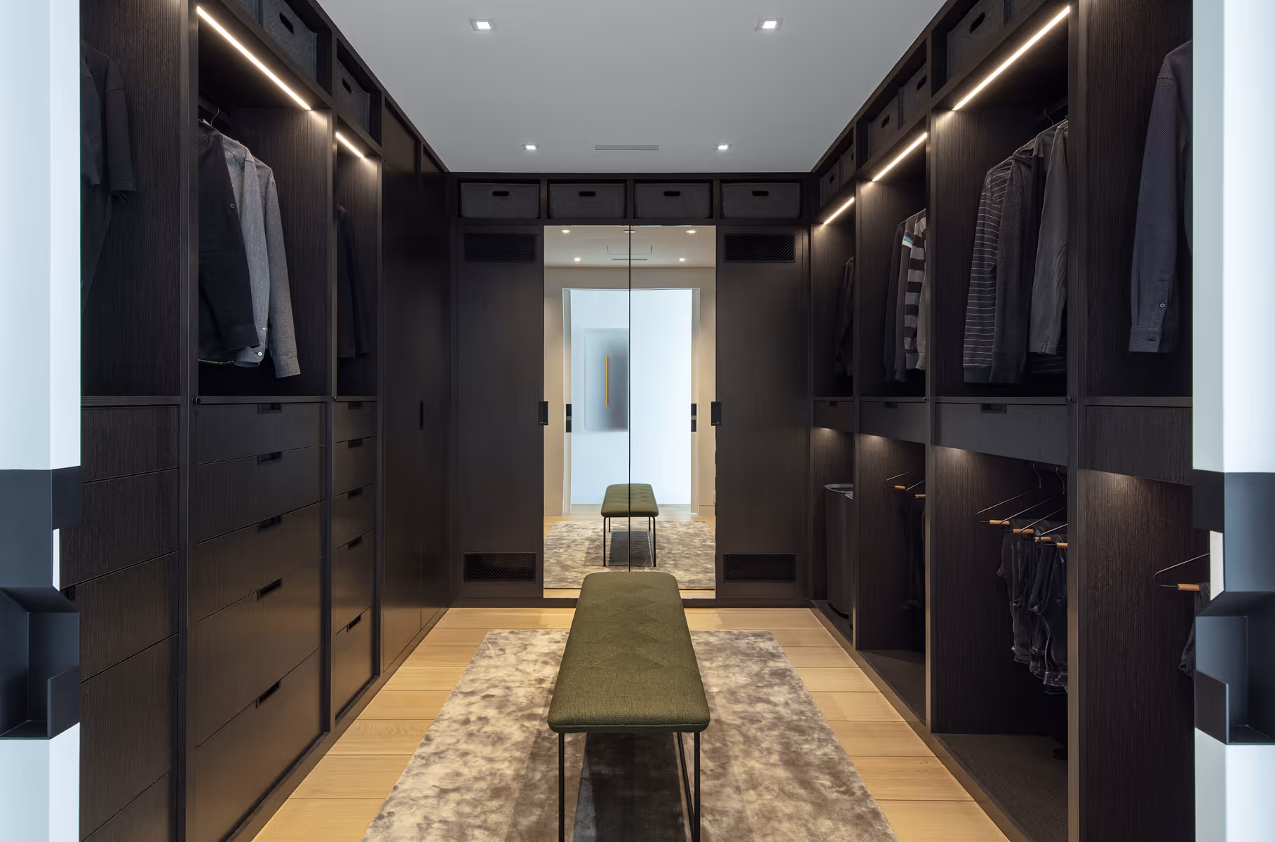 Modern walk-in closet with dark wood shelves, clothes, overhead lights, and green bench on rug.