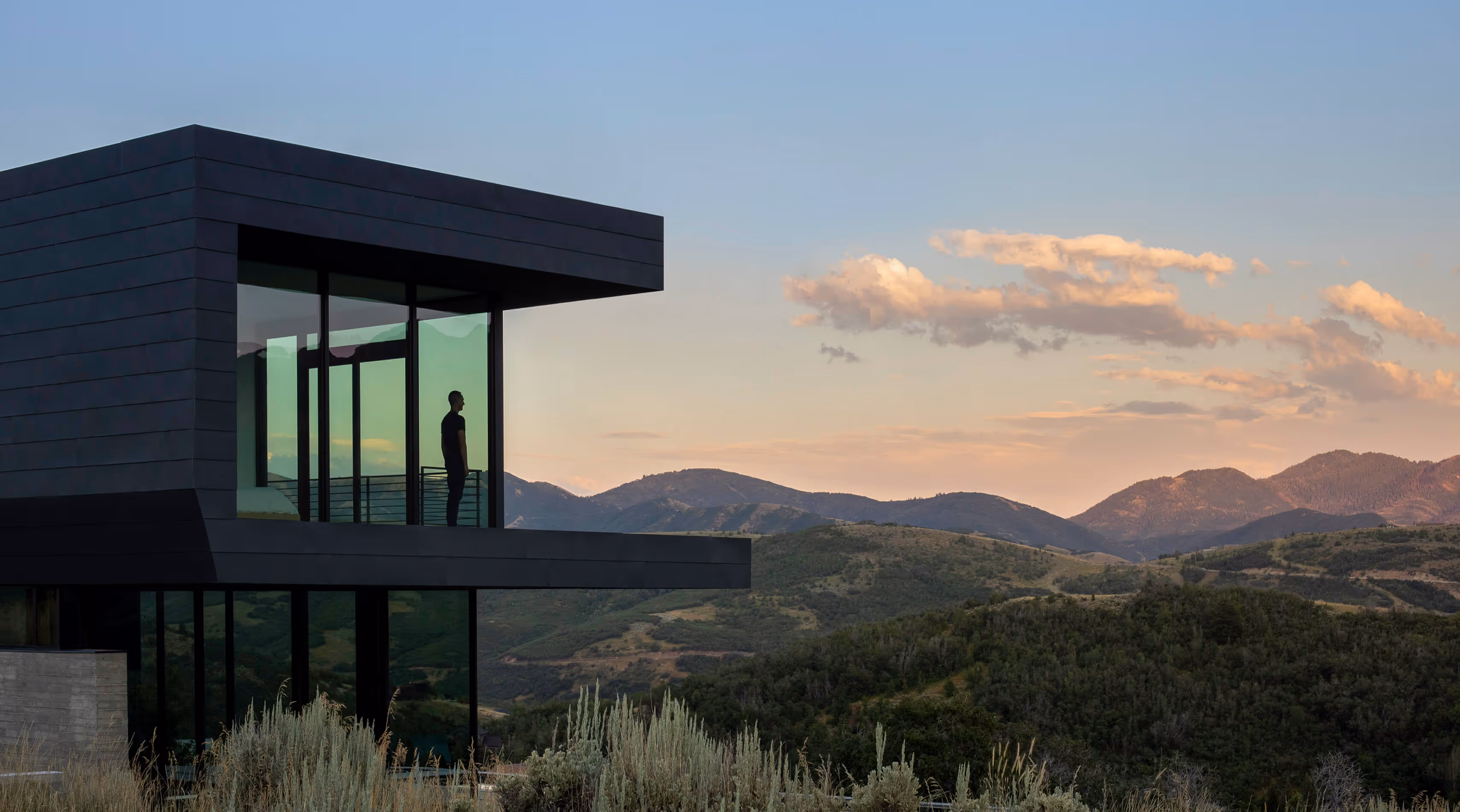 Person standing inside modern house with glass walls overlooking mountain landscape at sunset.
