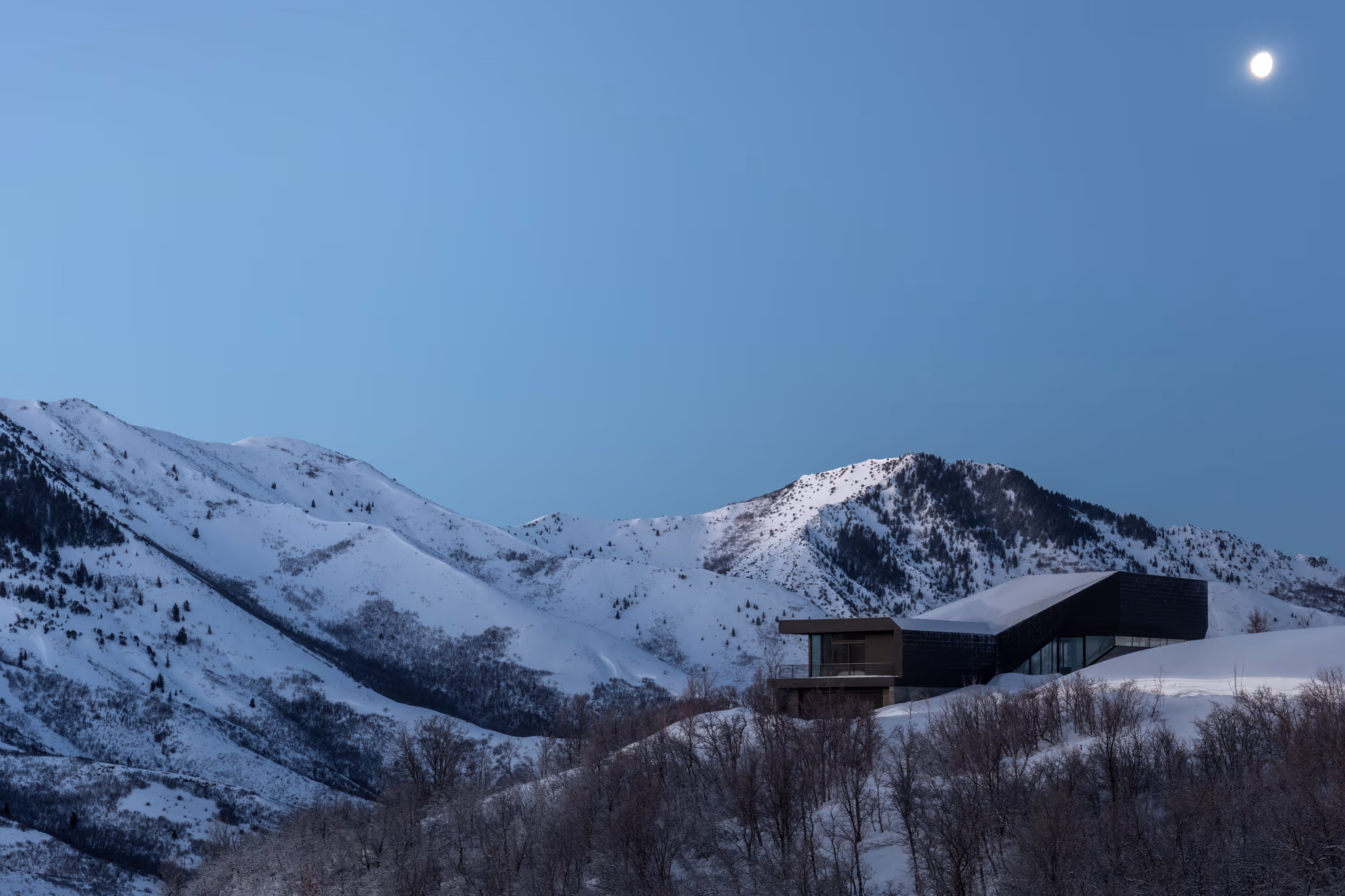 Modern black house on snowy hill with leafless trees and snow-covered mountains under moonlight.