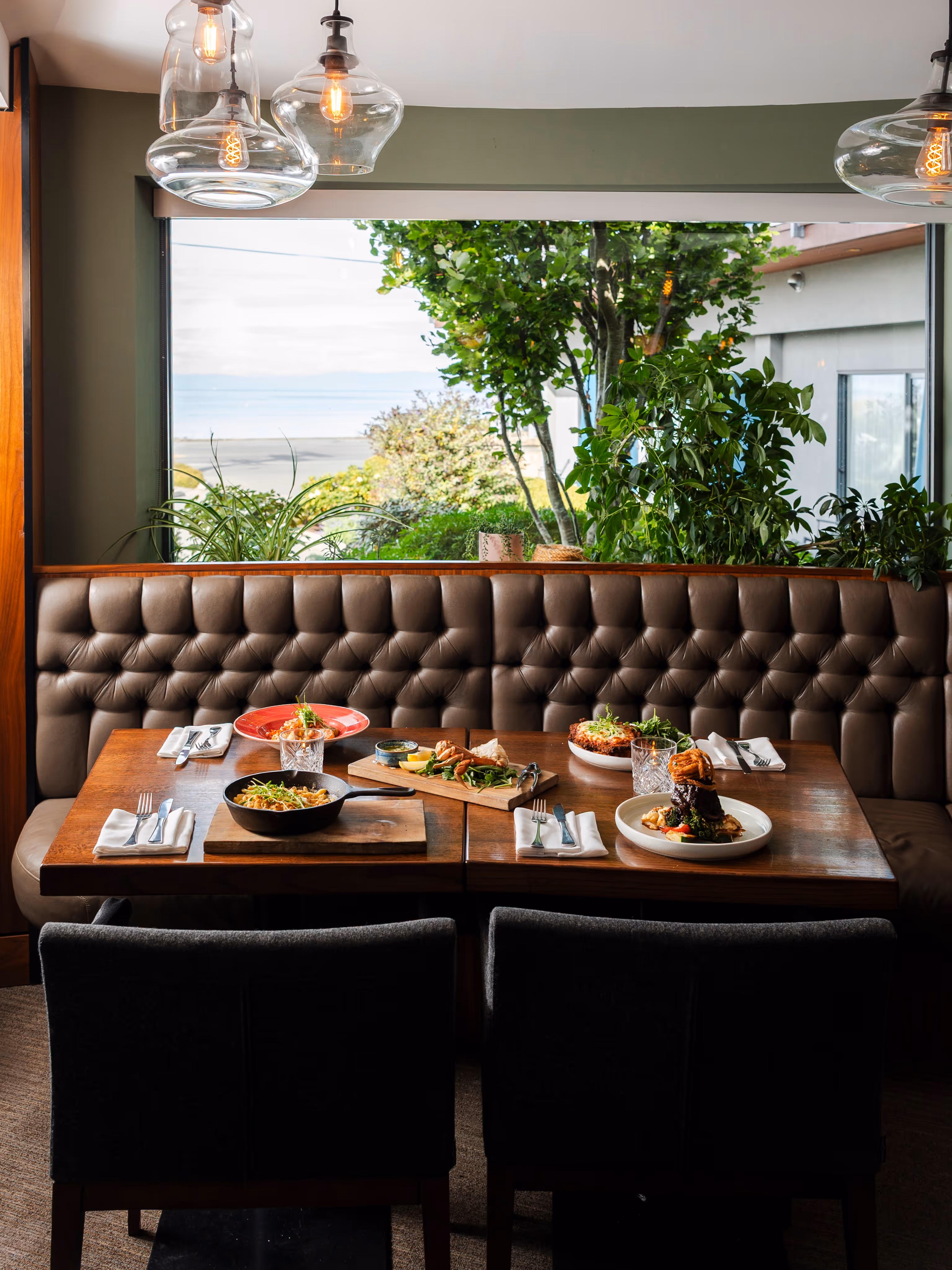 Restaurant dining table set with four plates of gourmet food, cutlery, and glasses, with a tufted leather bench and large window showing greenery outside.