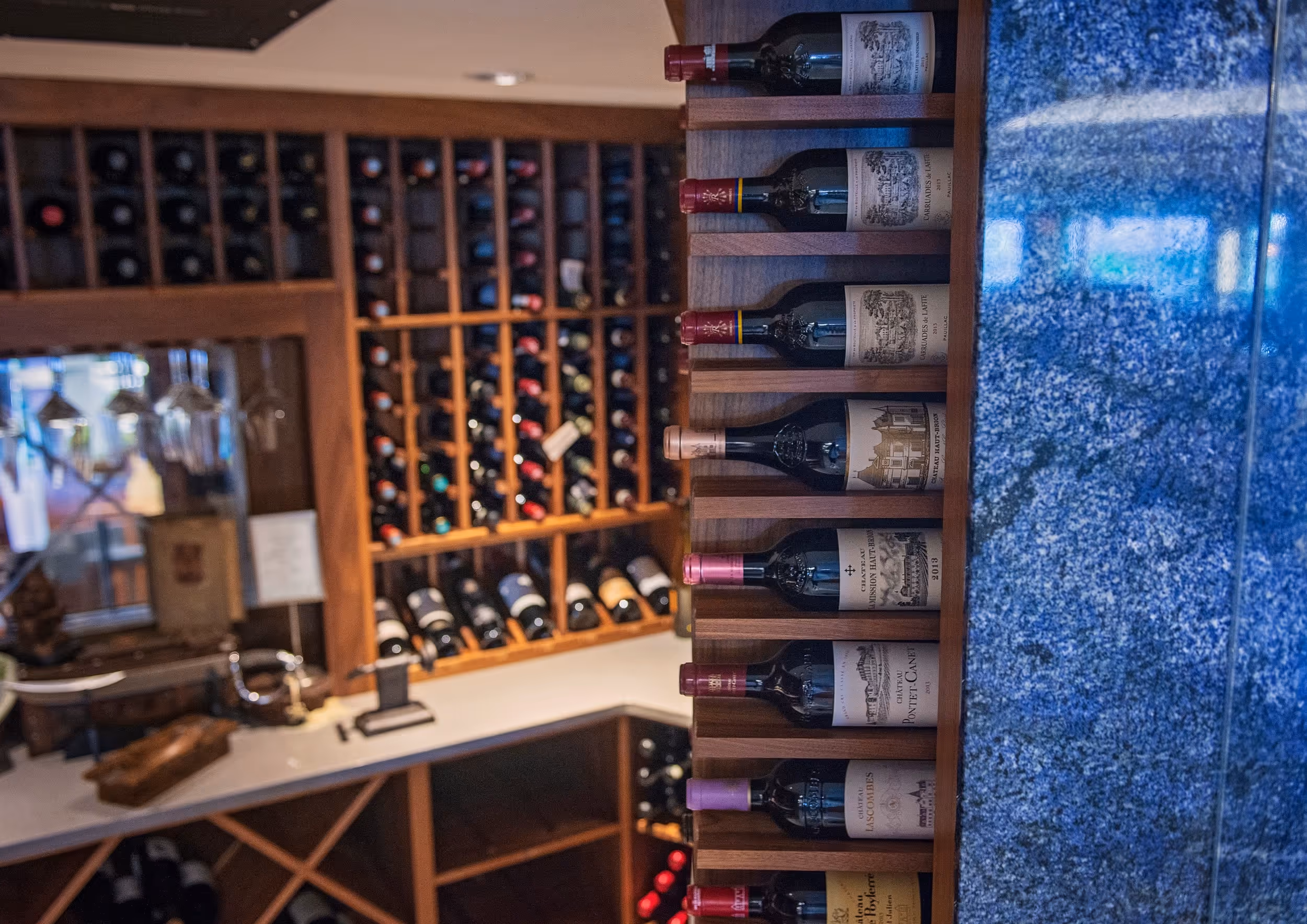 Shelves with seven bottles of red wine on the right and a wine storage rack filled with bottles in the background on the left.