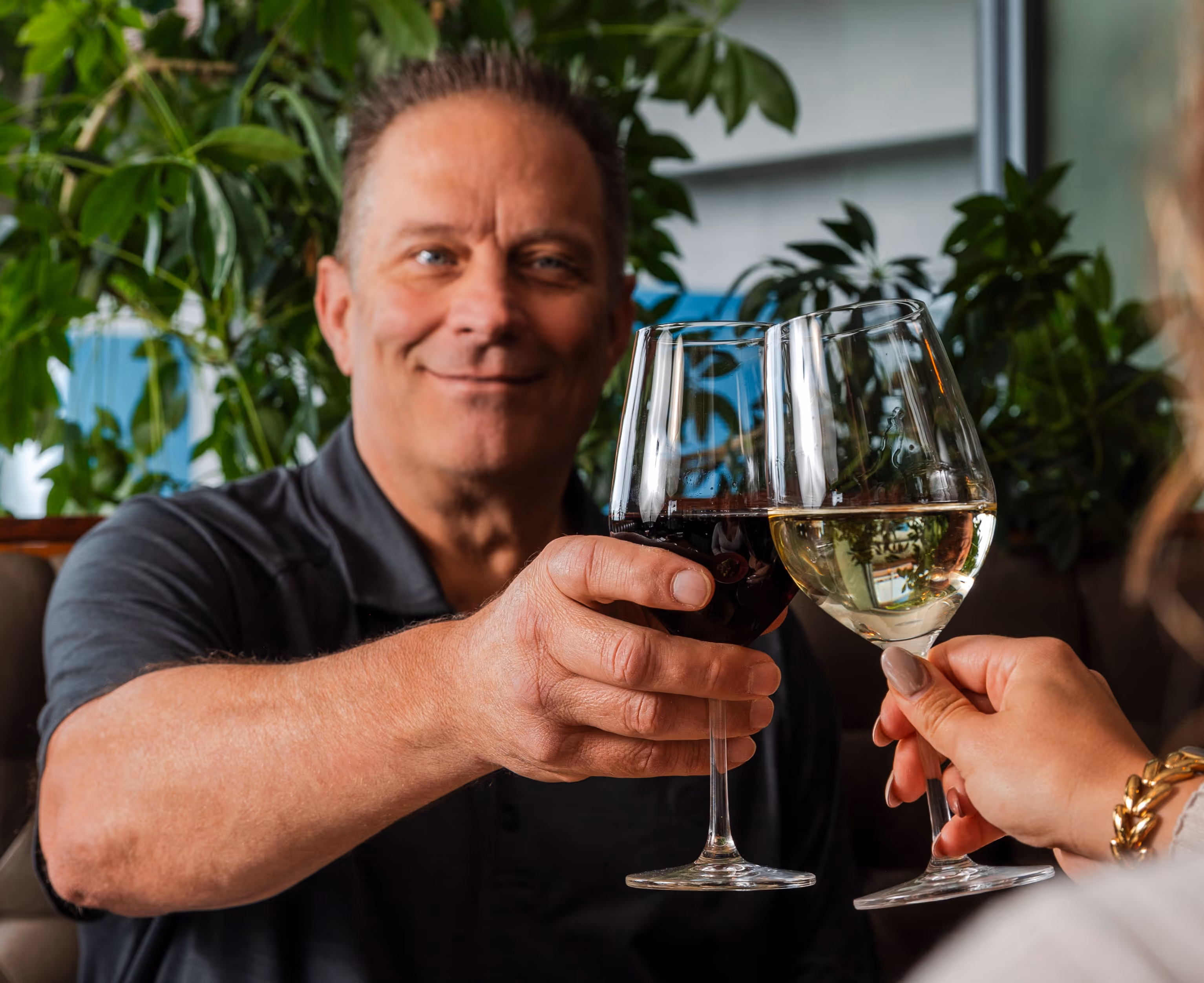 Man and woman clinking glasses of red and white wine in a restaurant with green plants in the background.
