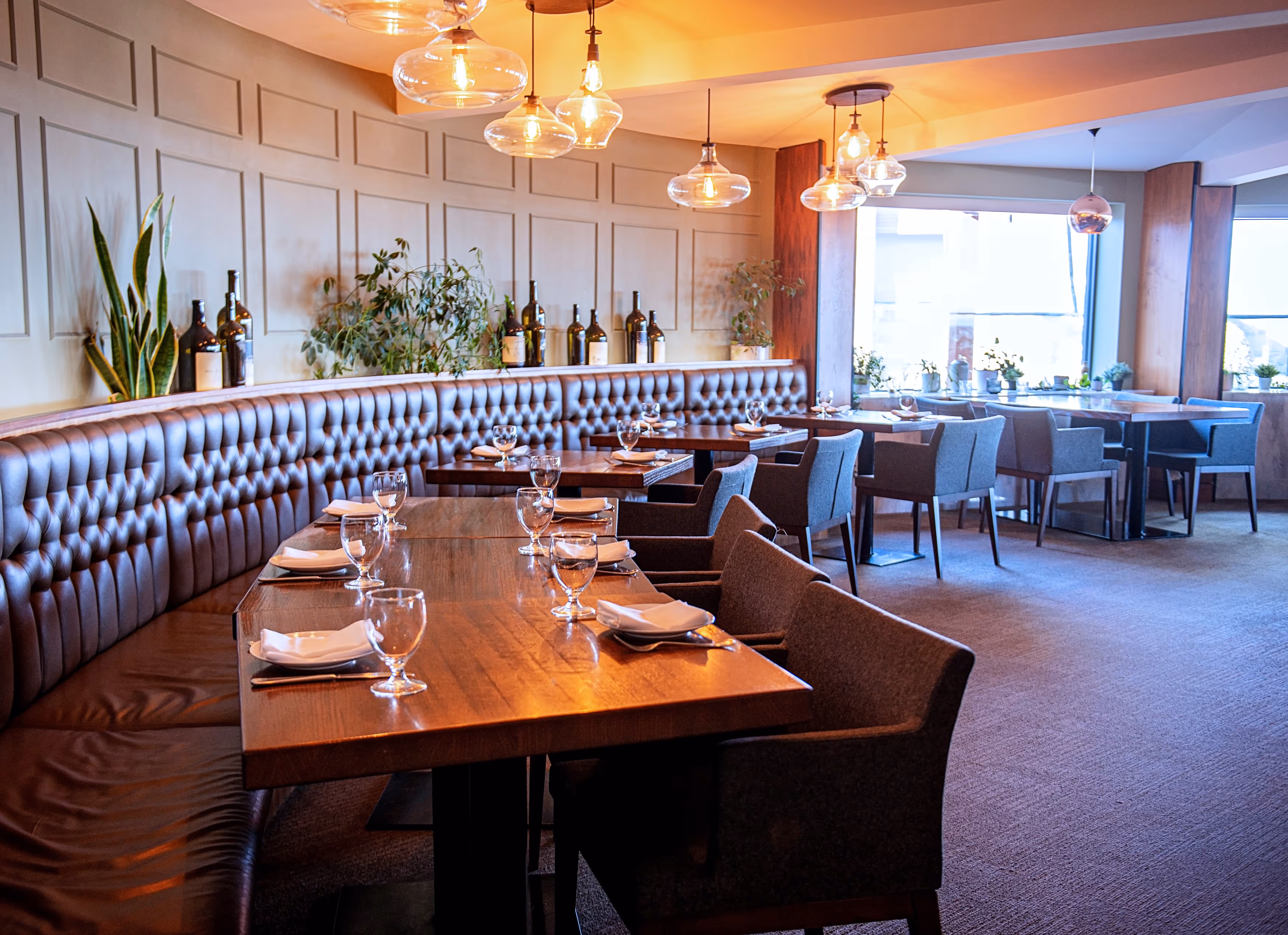 Empty restaurant dining area with wooden tables set with glasses and napkins, surrounded by padded chairs and a leather banquette along the wall, with hanging glass pendant lights and plants.