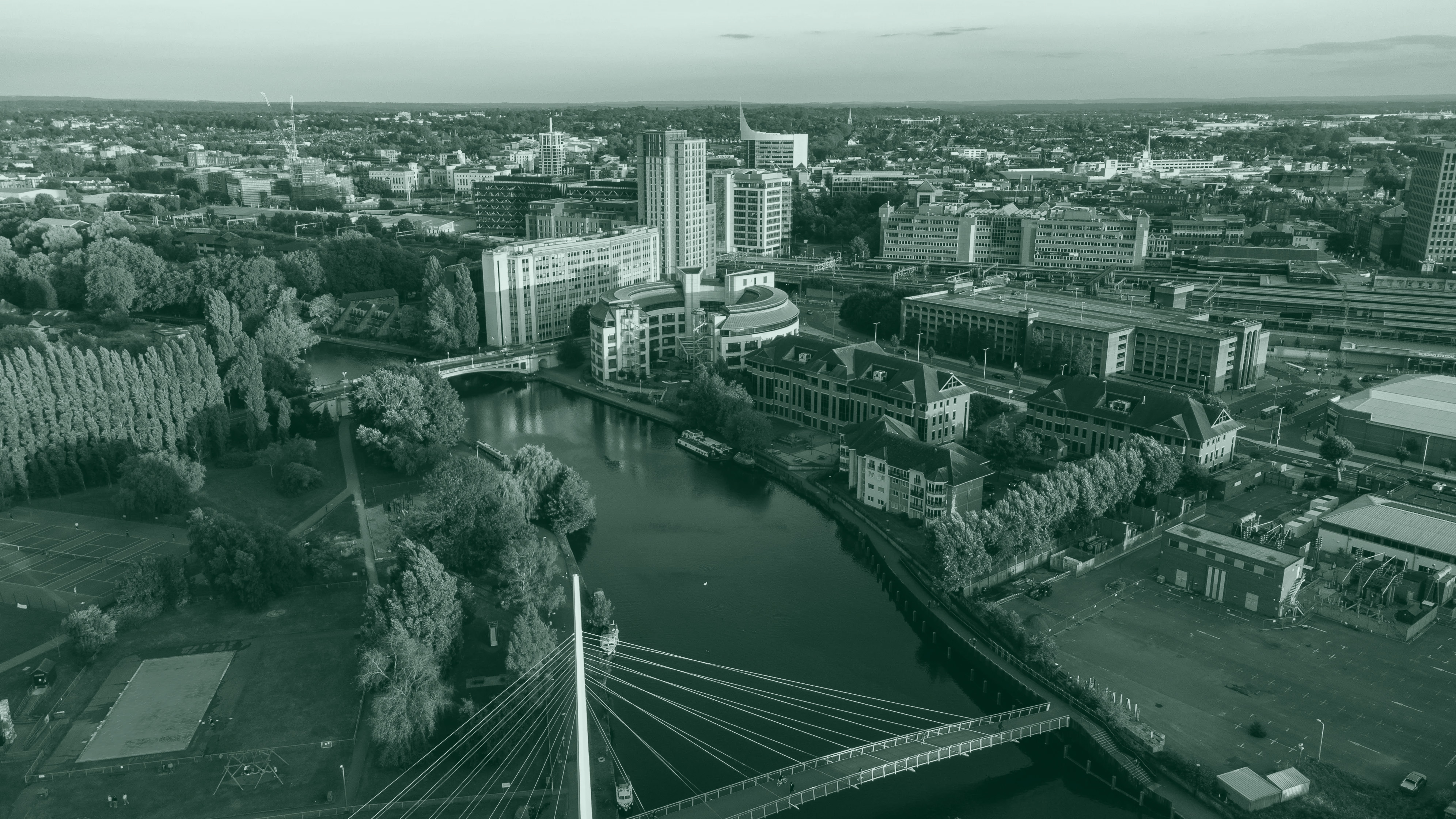 An aerial view of a city landscape during golden hour, featuring a river, a modern cable-stayed pedestrian bridge, and a mix of commercial buildings and green parks.