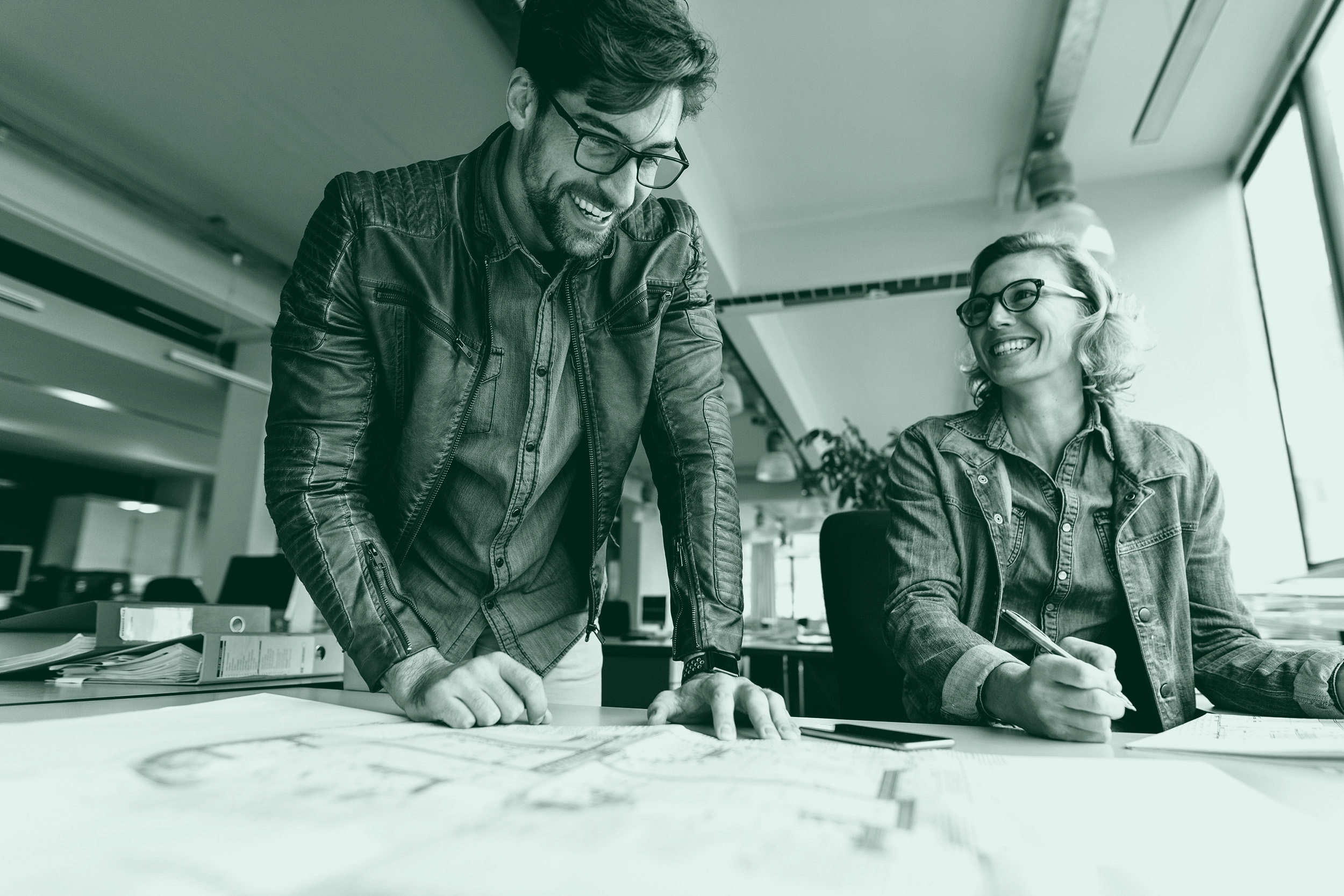 Two colleagues, a man and a woman, smiling and collaborating over large blueprints at a desk in a bright, modern office.