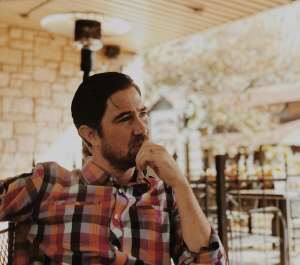 A thoughtful man in a plaid shirt sits at an outdoor café, gazing contemplatively to his left with his hand resting on his chin.