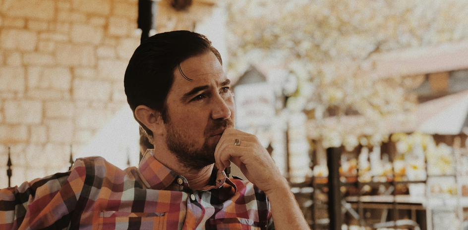 A thoughtful man in a plaid shirt sits at an outdoor café, gazing contemplatively to his left with his hand resting on his chin.