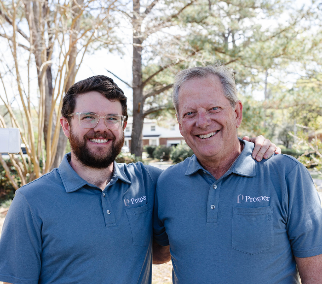 Three generations of a family, including a young man, an older man, and a toddler, smiling near a 'For Rent' sign from Prosper Property Management, set in a residential area with trees and a landscaped background.