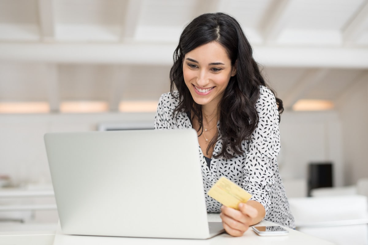 A joyful woman with dark hair shopping online, holding a credit card in hand and looking at her laptop screen, positioned in a bright office space, exemplifying convenience and the ease of digital transactions.