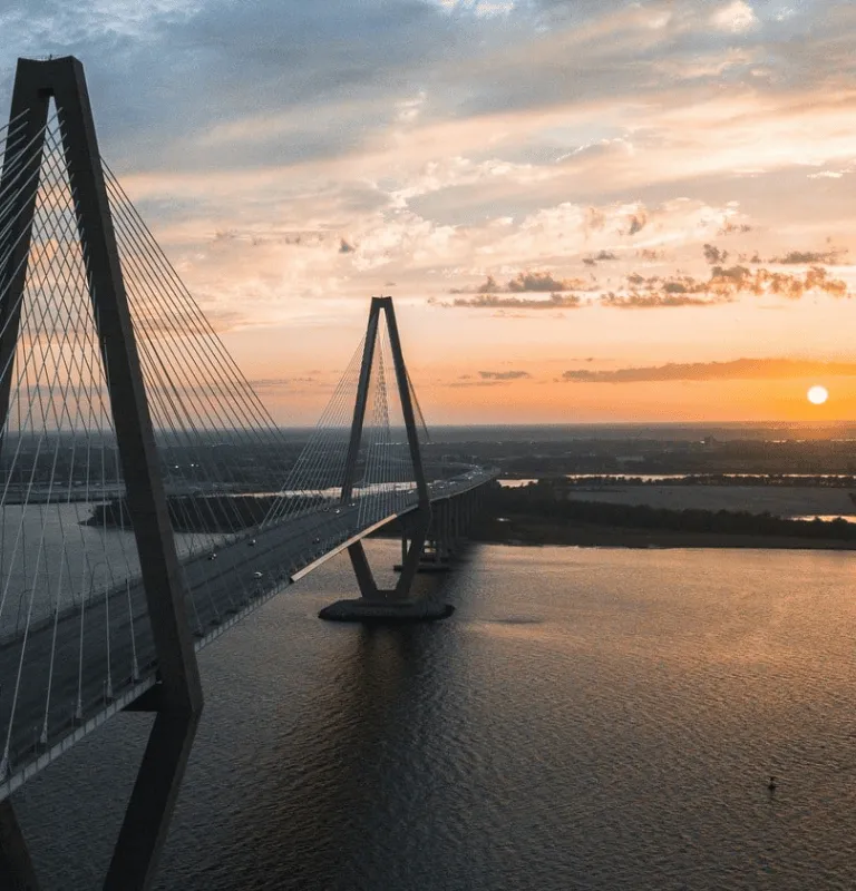 Sunset view of the Arthur Ravenel Jr. Bridge in Charleston, with a radiant sun dipping below the horizon, reflecting over the water.