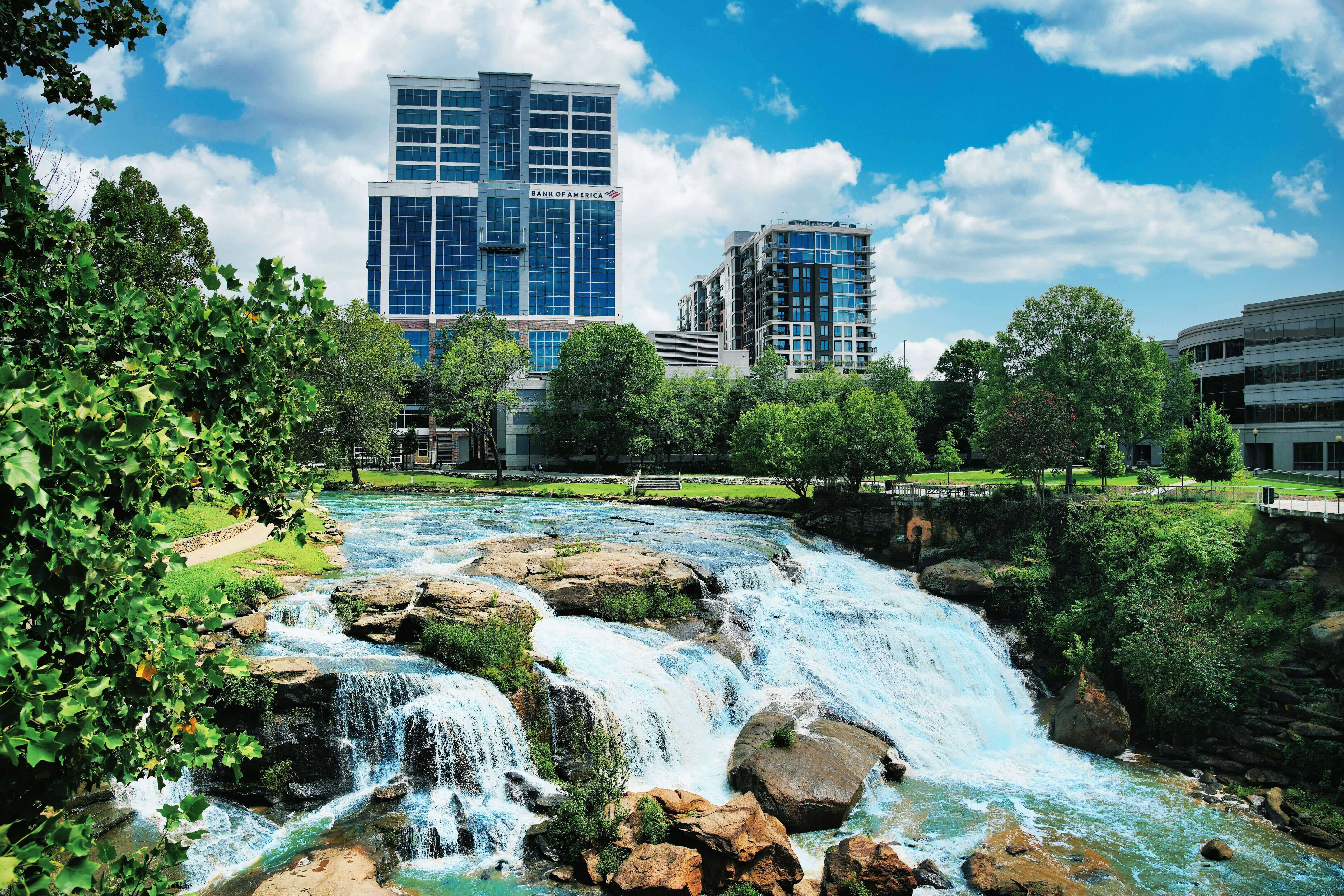 Waterfall cascading over rocks in an urban park with trees and modern buildings, including a Bank of America building, under a blue sky.
