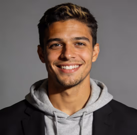 Smiling young man with short dark hair and blonde highlights wearing a gray hoodie and black blazer against a gray background.