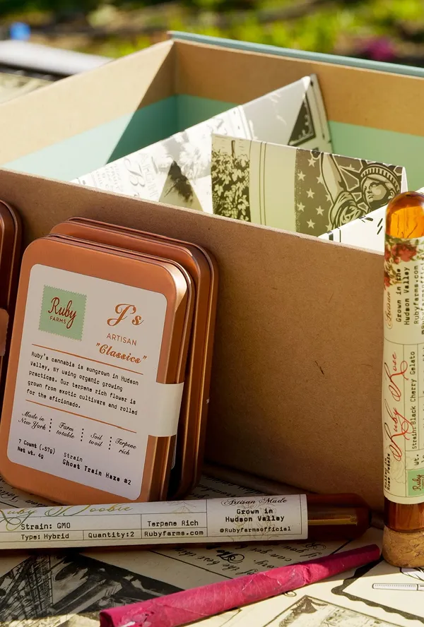 Copper tins labeled Ruby Farms cannabis products and tubes with strain info next to a cardboard box.