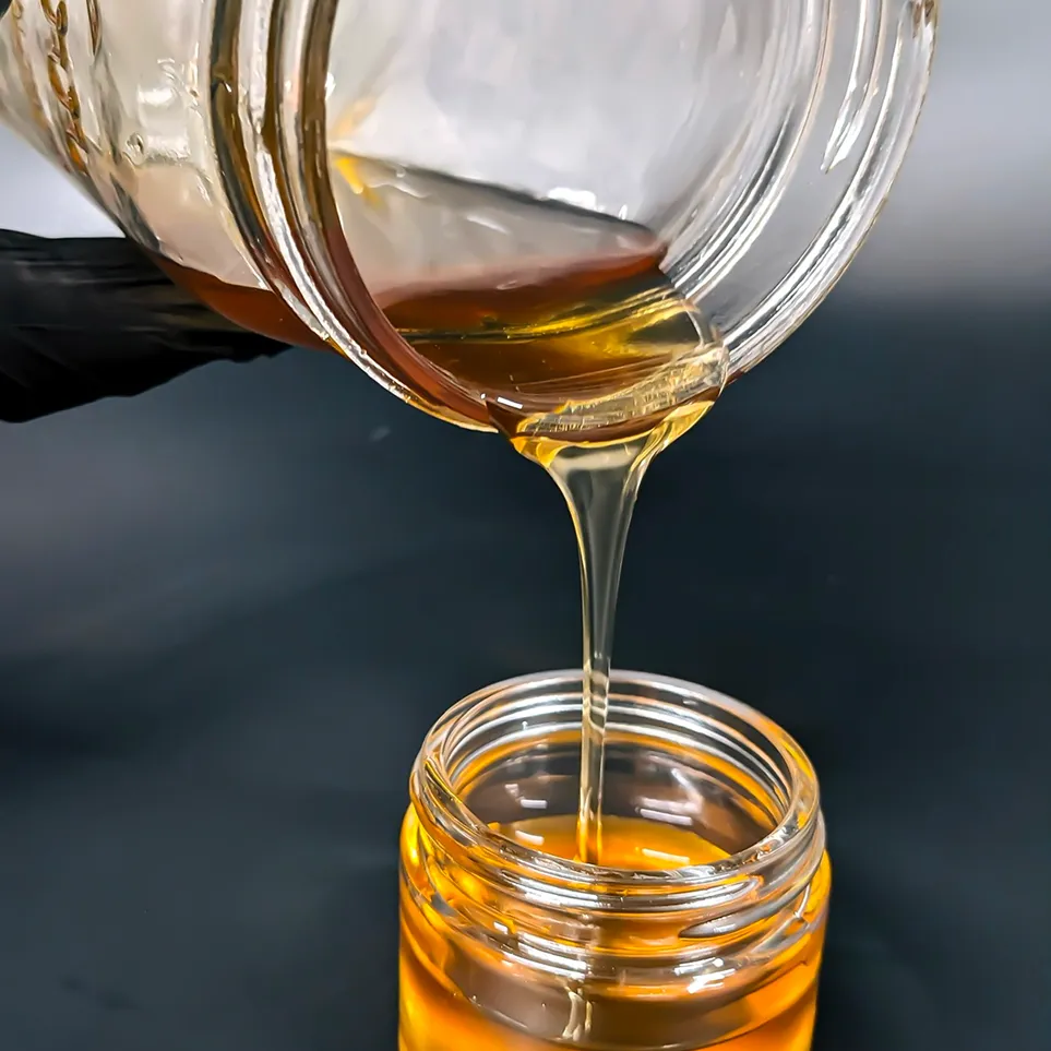 Golden honey pouring from a glass jar into another jar held by a hand wearing a black glove.
