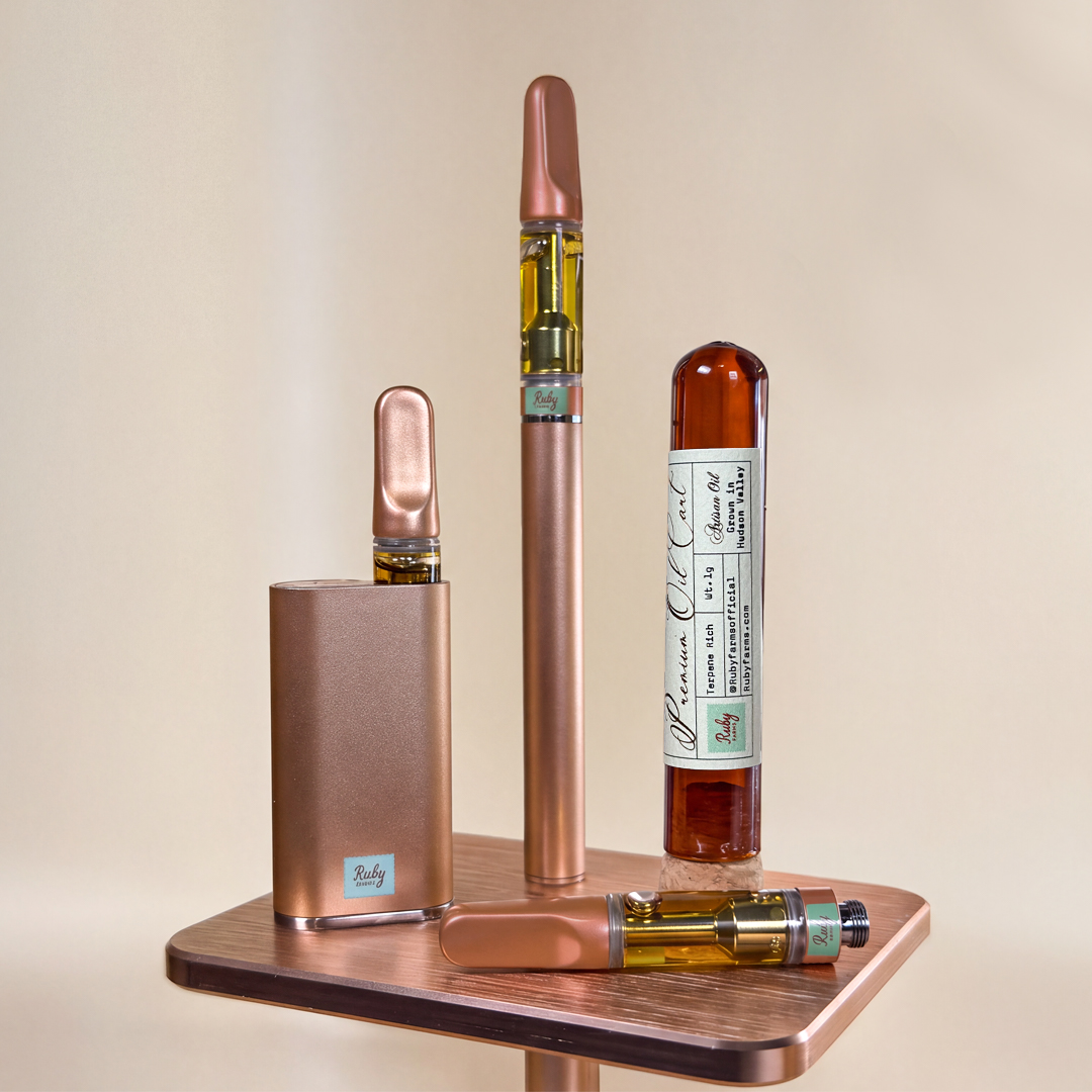 Bronze vape pens and cartridge with amber oil, labeled Ruby Farms, on wooden surface with forest background.
