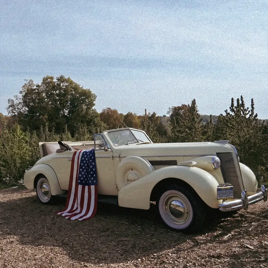 Vintage cream convertible car parked near a wooden cart with a white cloth cover outdoors.