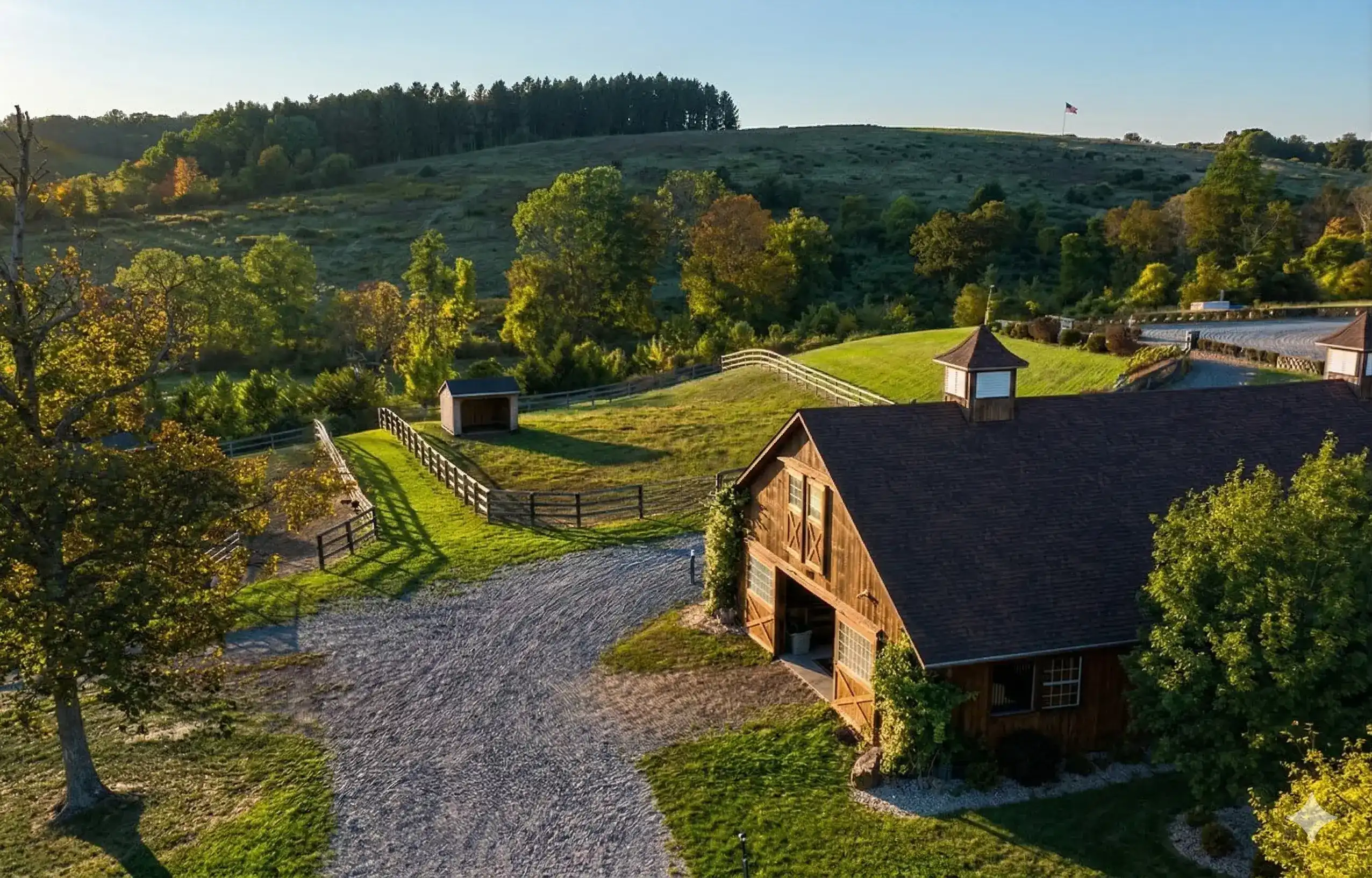 Wooden barn beside a gravel driveway with fenced pasture and trees on rolling green hills.