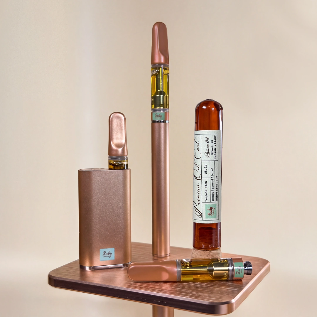 Bronze vape pens and cartridge with amber oil, labeled Ruby Farms, on wooden surface with forest background.