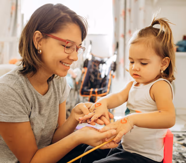 Young woman painting red hearts on a toddler girl's hands while both sit indoors.