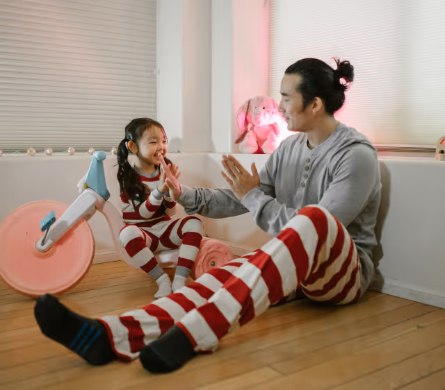 Father and daughter in matching red and white striped pajamas playing and clapping hands on a wooden floor near a pink toy bike indoors.