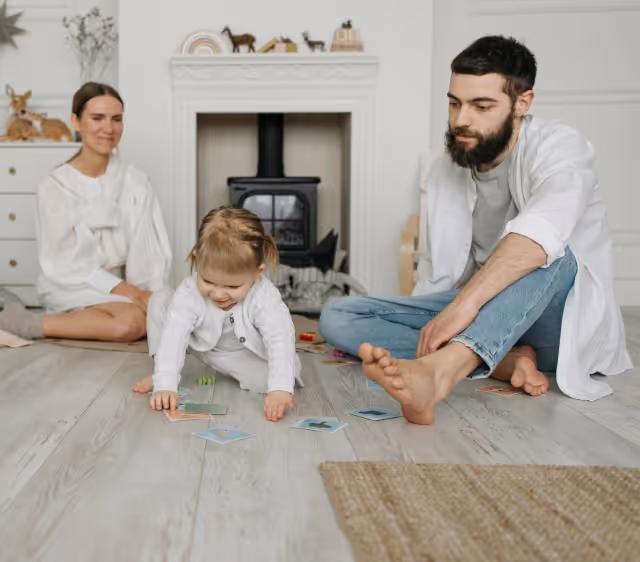 A toddler playing with cards on the floor while a man and a woman watch in a cozy living room with a fireplace.