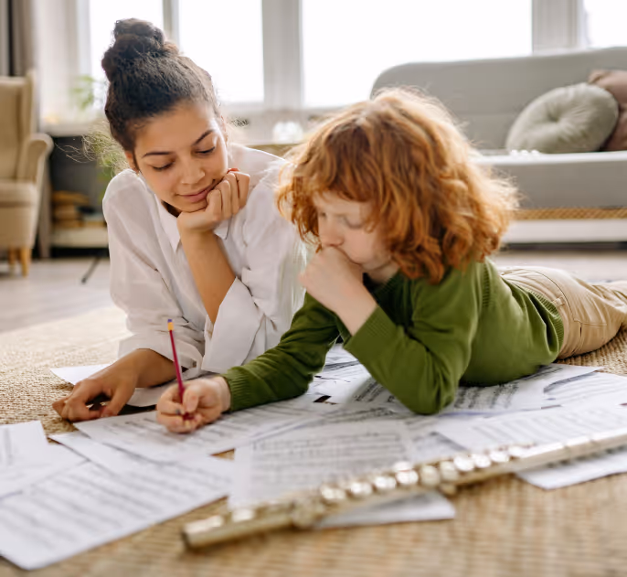 A woman and a child lying on the floor surrounded by sheet music, with a flute nearby, practicing music together.