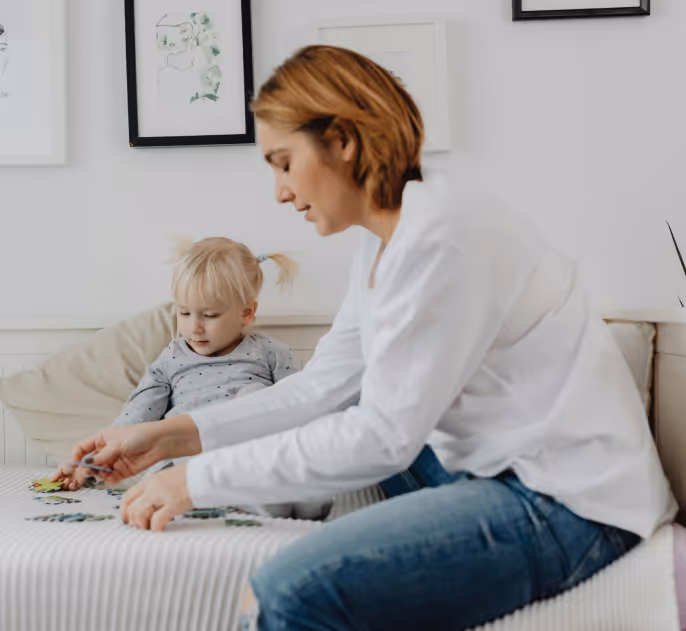 Woman and young child sitting on a bed working together on a puzzle.