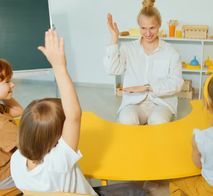 Teacher and children sitting around a yellow table in a classroom, raising their hands.