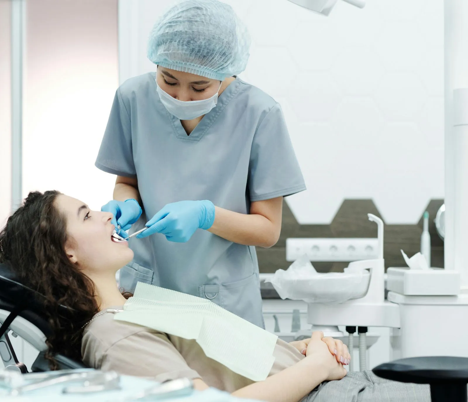 Dentist wearing scrubs, mask, and hair cover examining a female patient's teeth with dental tools in a clinic.