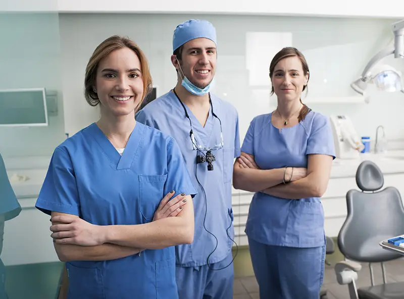 Three smiling dental professionals in blue scrubs standing confidently in a dental clinic.