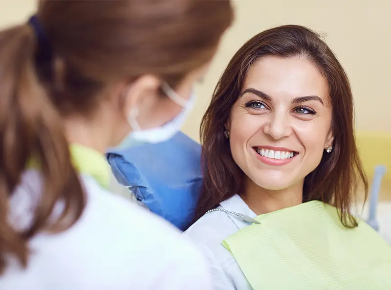 Smiling woman sitting in dental chair talking to female dentist wearing a face mask.