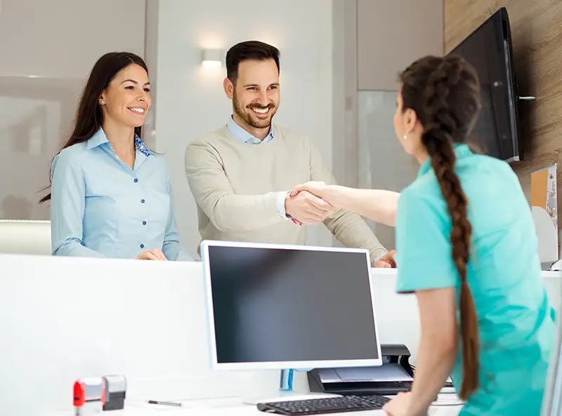 Man and woman smiling and shaking hands with a receptionist wearing teal scrubs at an office desk.