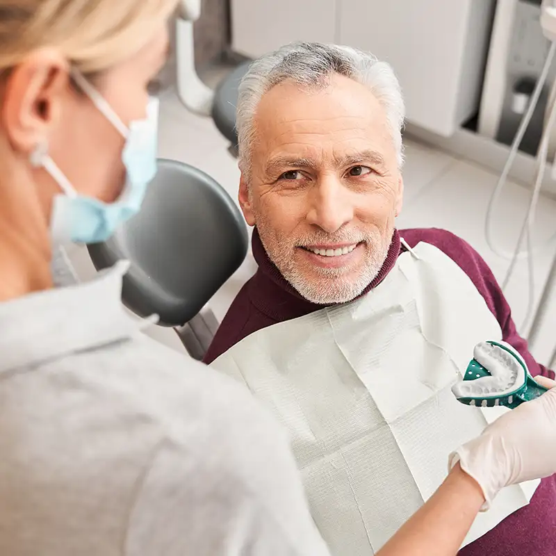 Smiling elderly man sitting in a dental chair looking at a masked dental professional holding a dental impression tray.