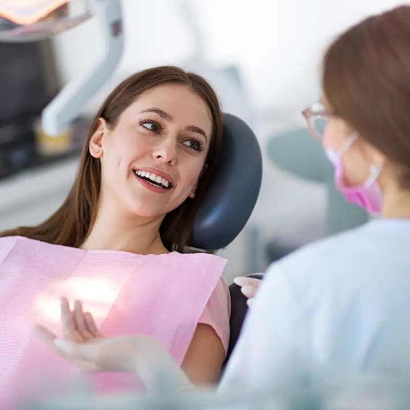 Smiling woman wearing a pink dental bib talking to a female dentist in a white coat and pink mask.