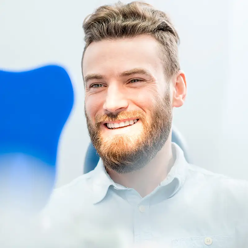 Smiling man with a beard and light-colored shirt sitting in a bright room.