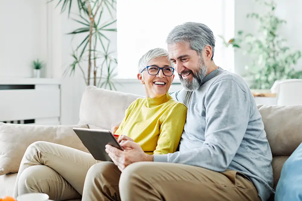 Smiling senior couple sitting on a couch looking at a tablet together in a bright living room.