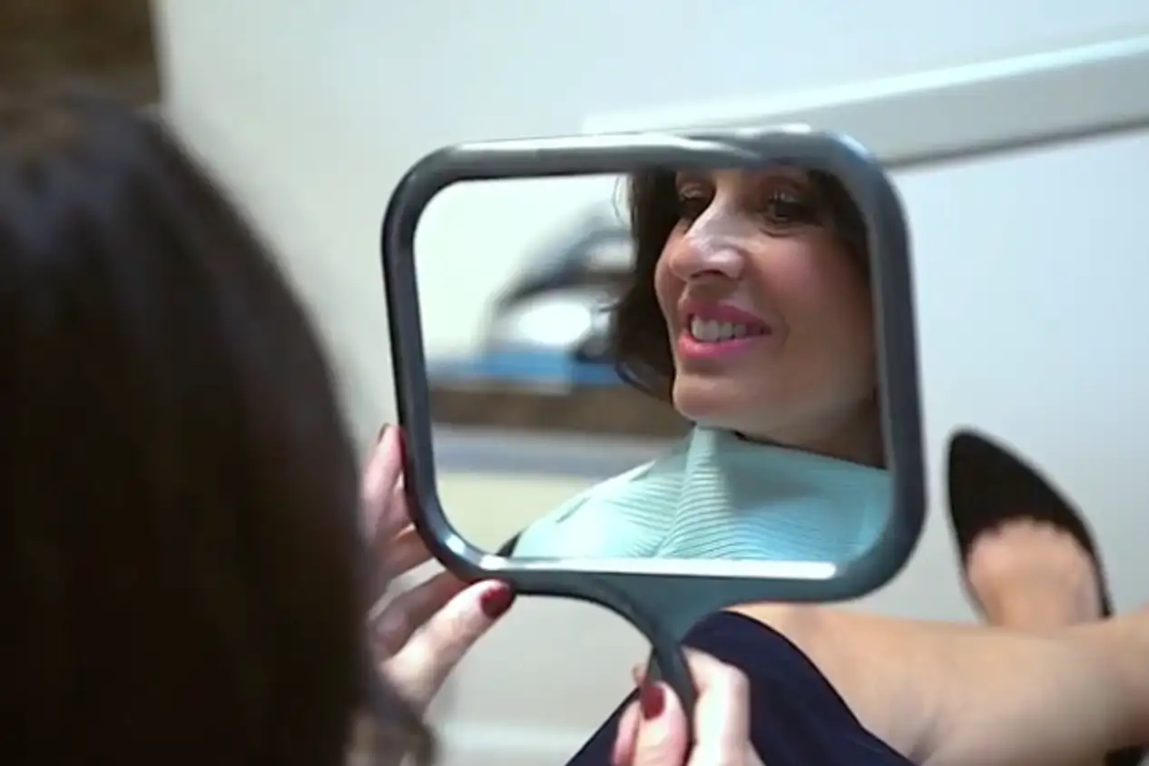 Woman smiling while looking at her reflection in a handheld mirror.